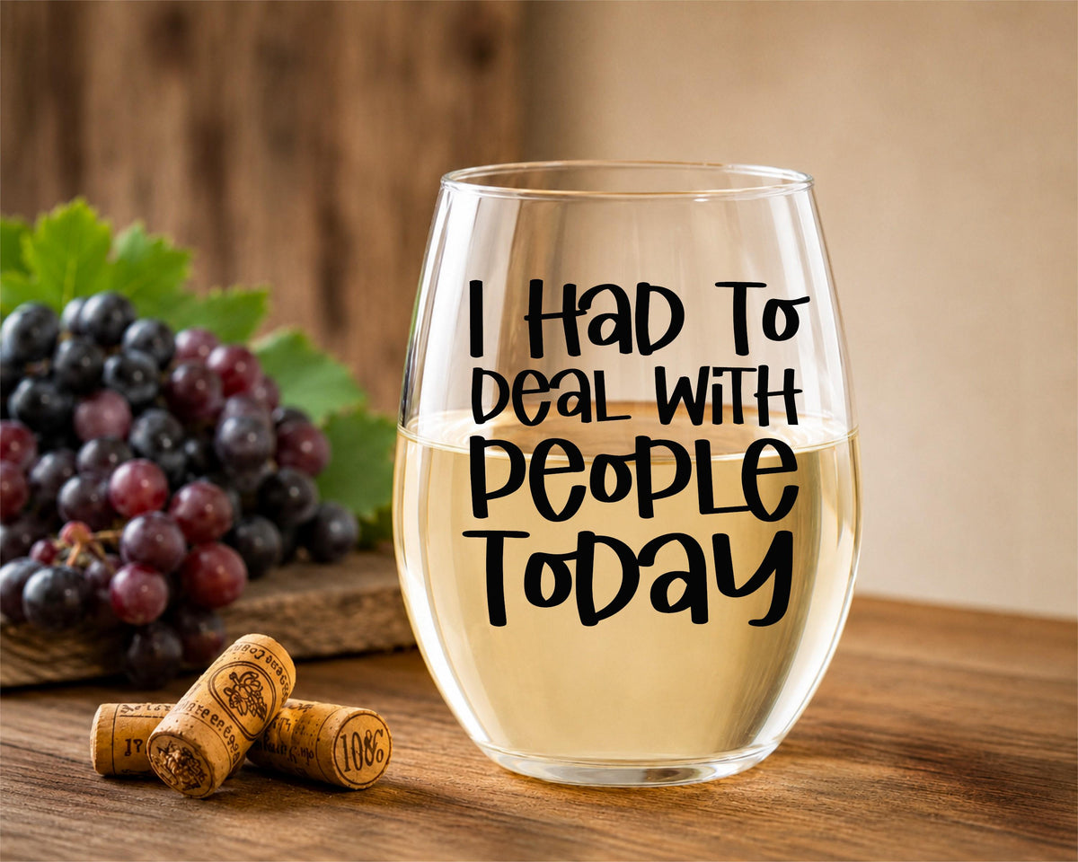 A stemless wine glass filled with white wine sits on a wooden table, printed with the phrase 'I HAD TO DEAL WITH PEOPLE TODAY' in black lettering. Grapes and wine corks are placed beside it, with a warm rustic background.